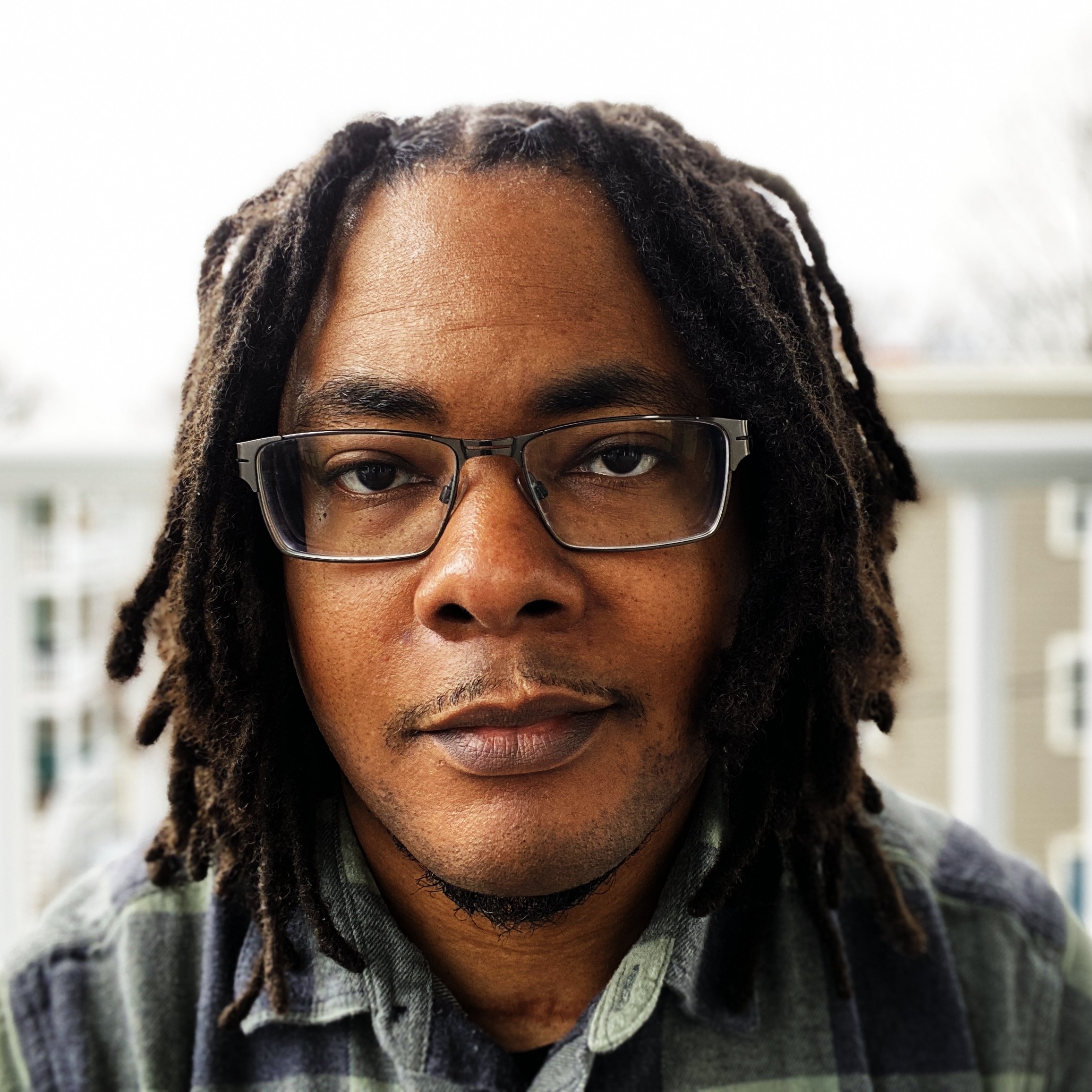 A smiling Black American man with long locs, glasses, and a button down shirt