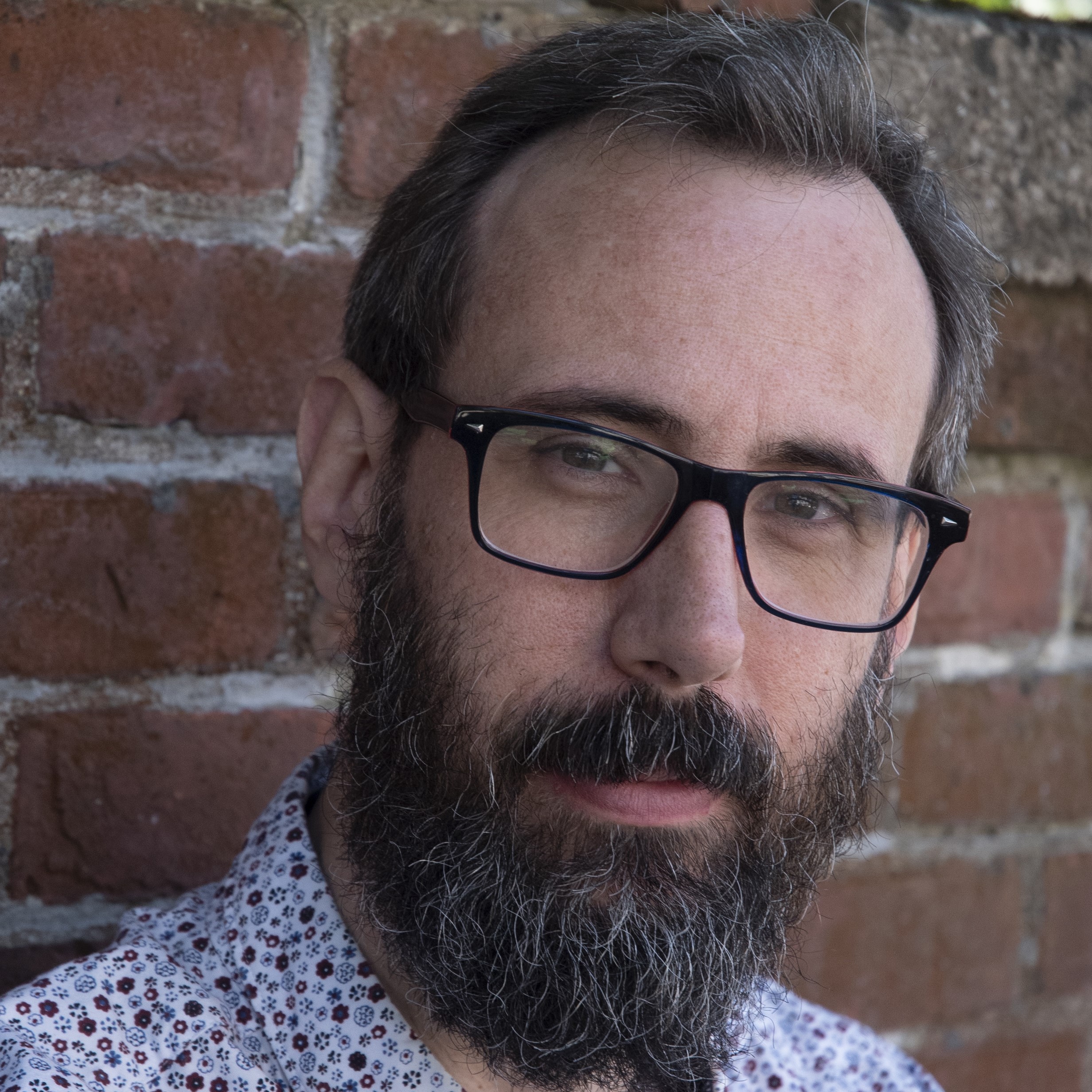 a white man with salt-and-pepper hair and beard and glasses in front of a brick wall