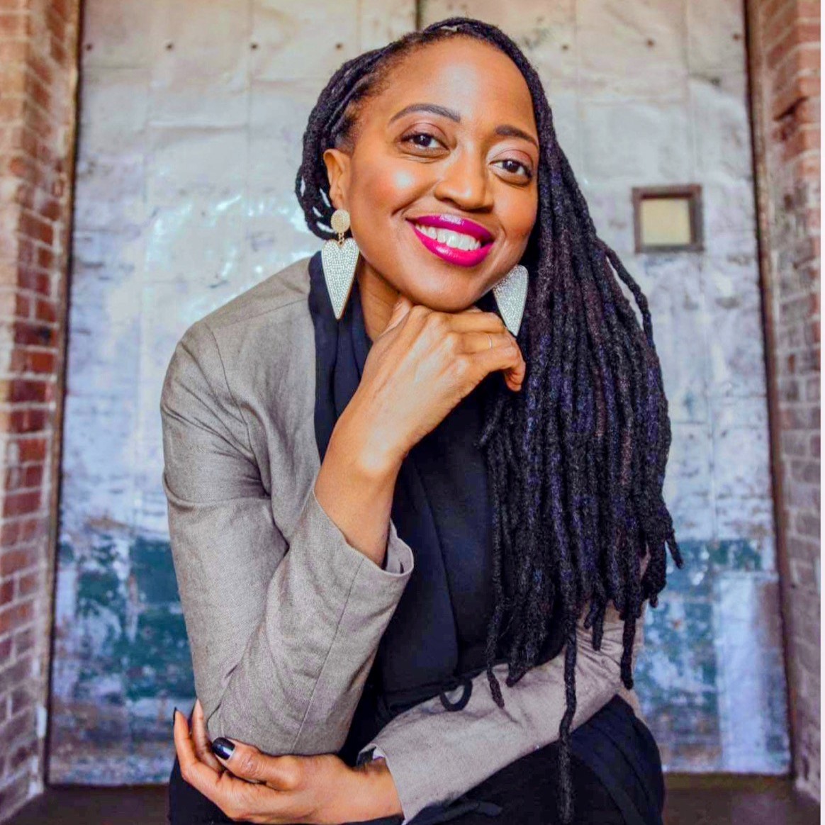 a smiling Black woman with heart-shaped earrings, long locs, blazer, and scarf in front of a steel door bordered by brick walls