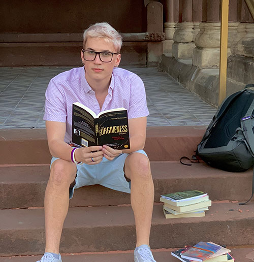 Male college student with blond hair and glasses, wearing pink shirt and blue short, sits on stairs looking up from reading a book